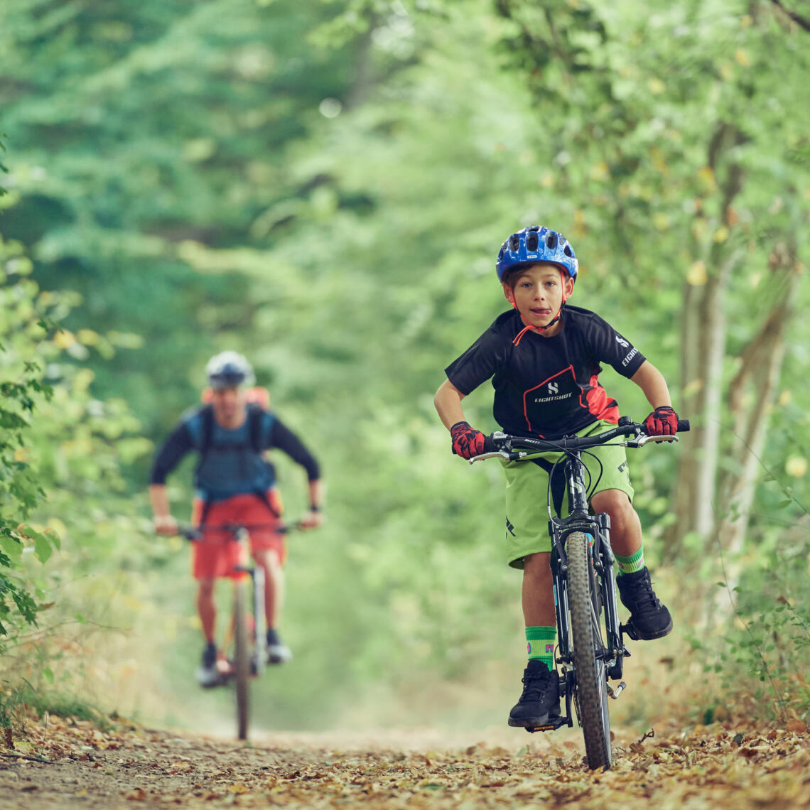 Junge fährt mit Mountainbike durch den Wald. Sein Vater fährt im Hintergrund