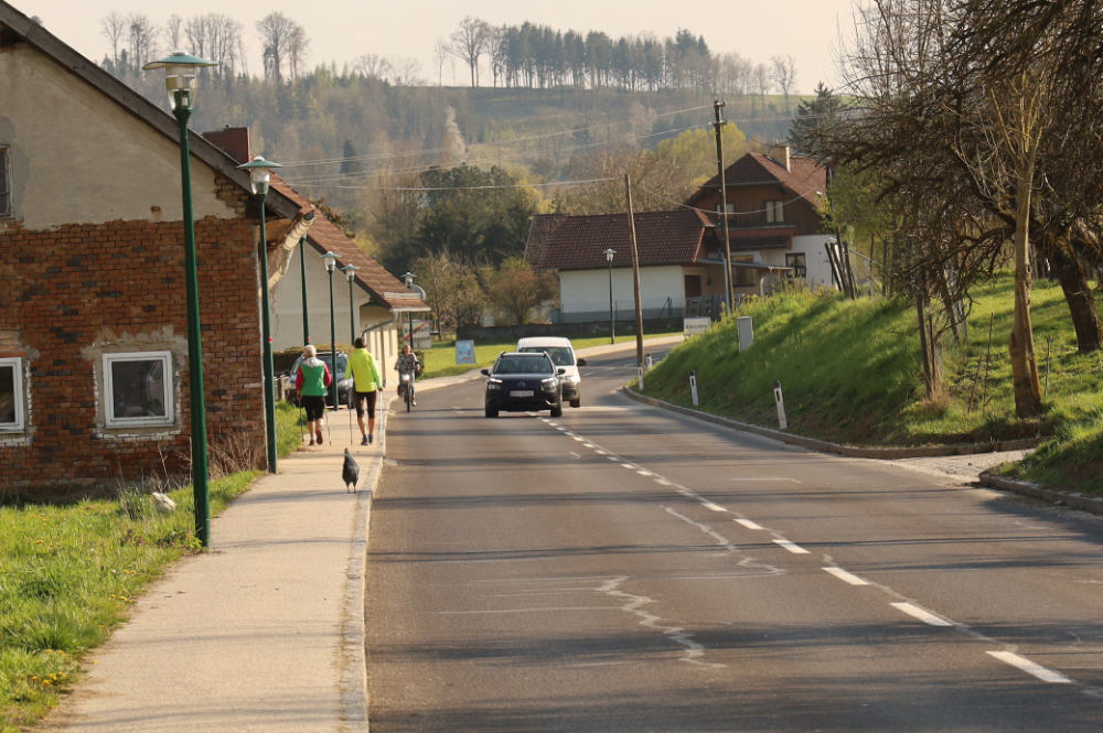 Landstraße im Ortsgebiet mit zwei Kfz, die eine Radfahrerin überholen, am Gehsteig daneben sind zwei Fußgängerinnen und ein Huhn