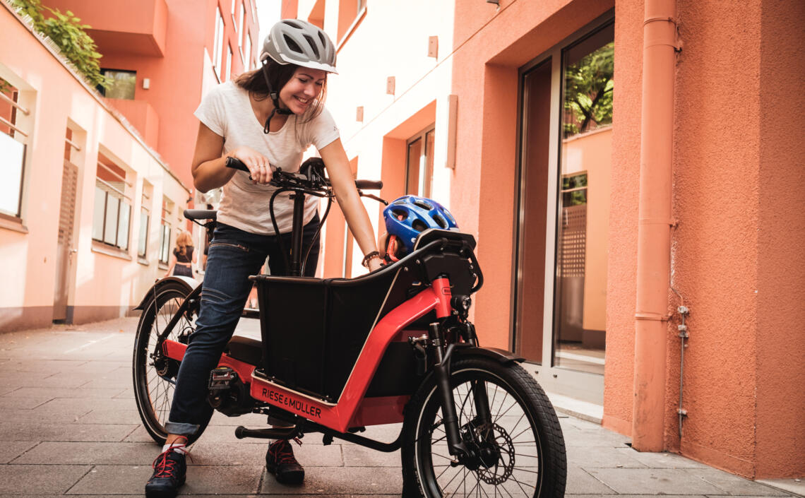 Dame steht mit Lasten-Fahrrad Riese und Müller und redet mit Kind, welches vorne im Korb sitzt