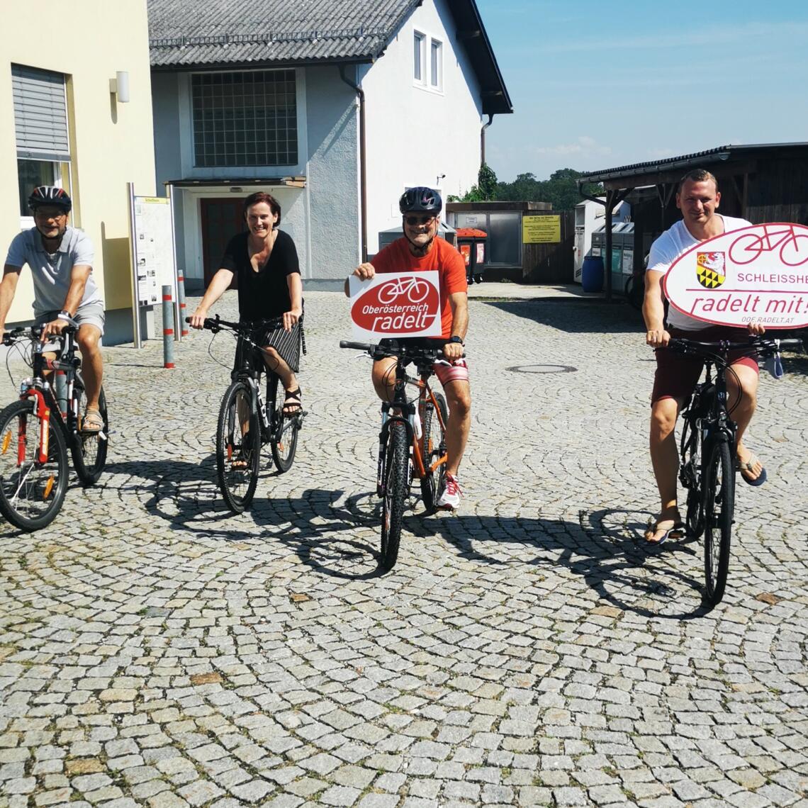 4 Erwachsene fahren mit dem Fahrrad und präsentieren Tafeln " Oberösterreich radelt" und "Schleissheim radelt mit"