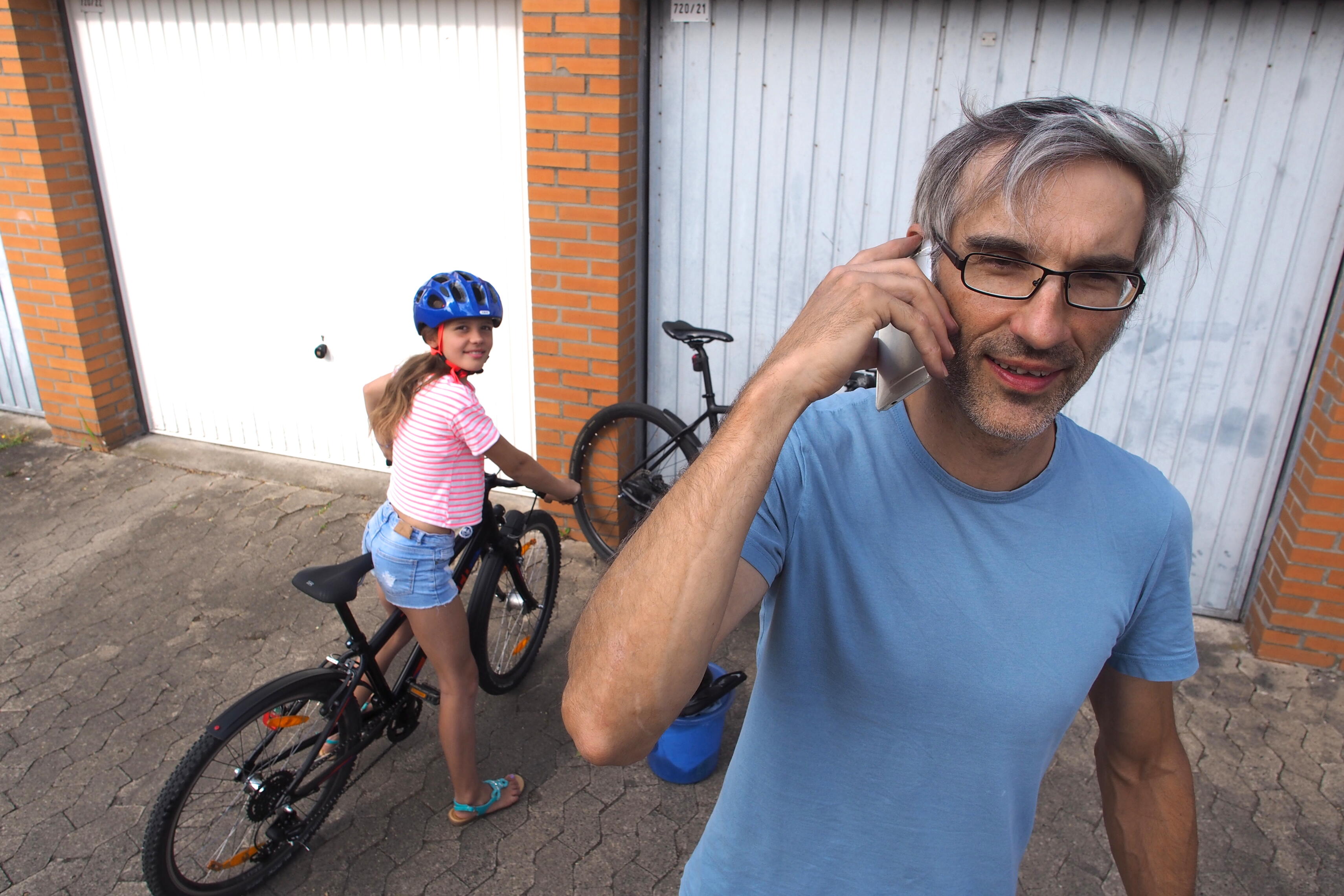 Mädchen steht mit Fahrrad vor Garagentor. Papa telefoniert mit Handy.