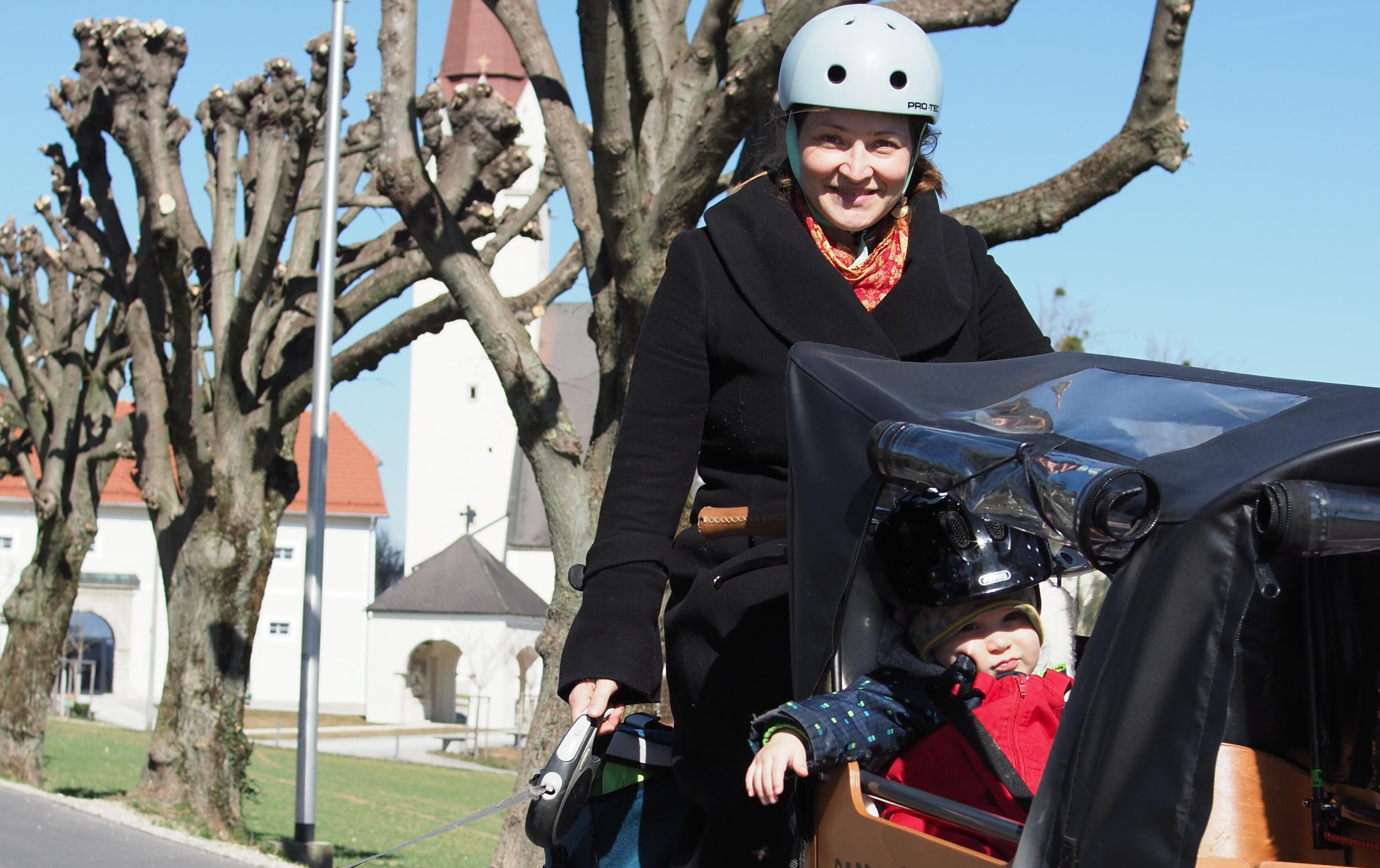 Dame fährt mit Lasten-Fahrrad. Im Aufsatz vor ihr sitzt ein Kind. Im Hintergrund ist eine Kirche erkennbar.