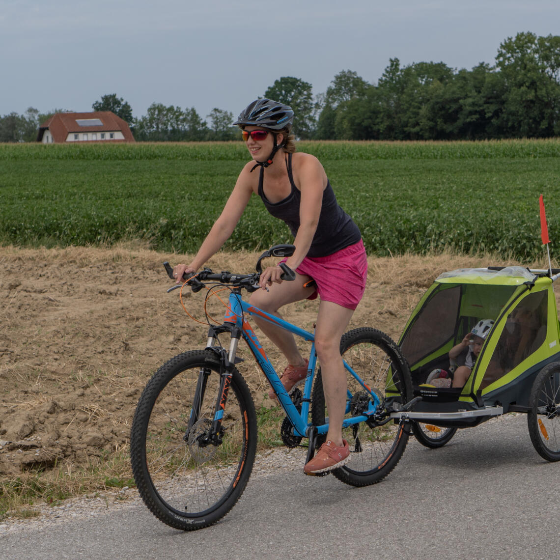 Dame fährt mit einem blauen Fahrrad mit Kinderanhänger auf der Straße neben einem Acker.