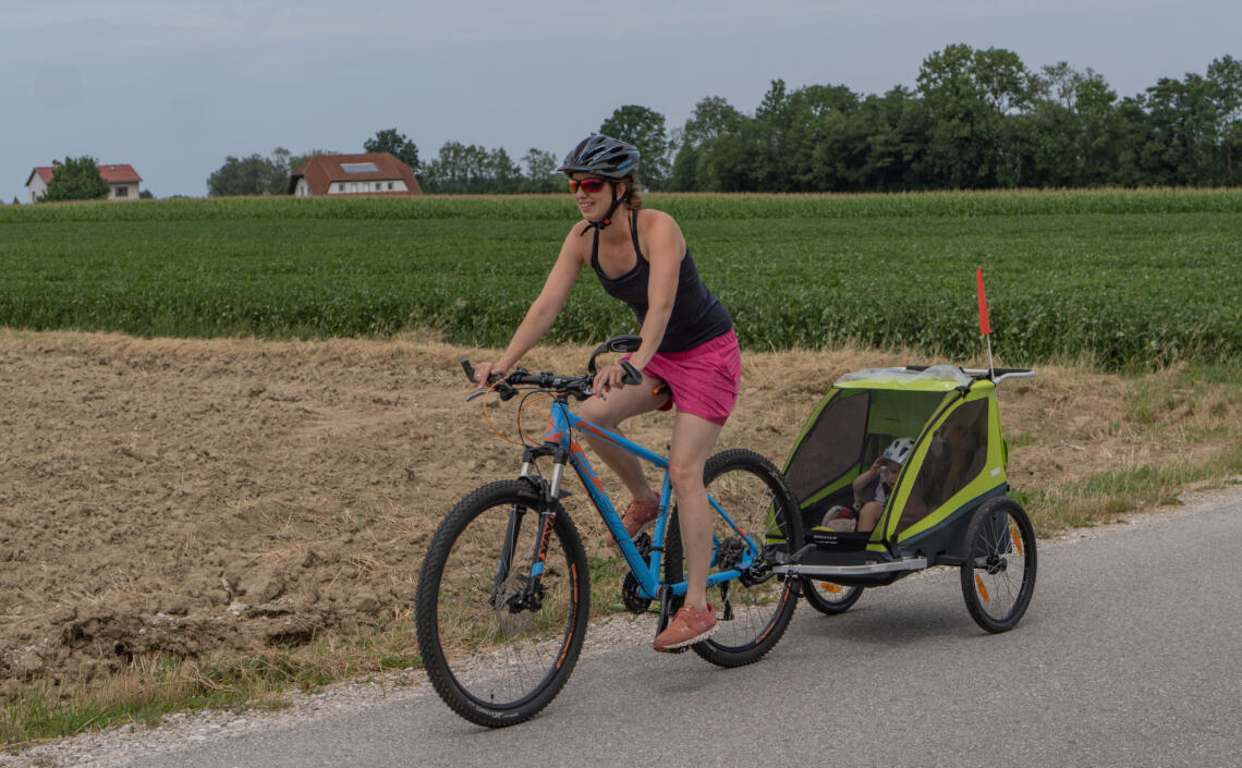 Dame fährt mit einem blauen Fahrrad mit Kinderanhänger auf der Straße neben einem Acker.