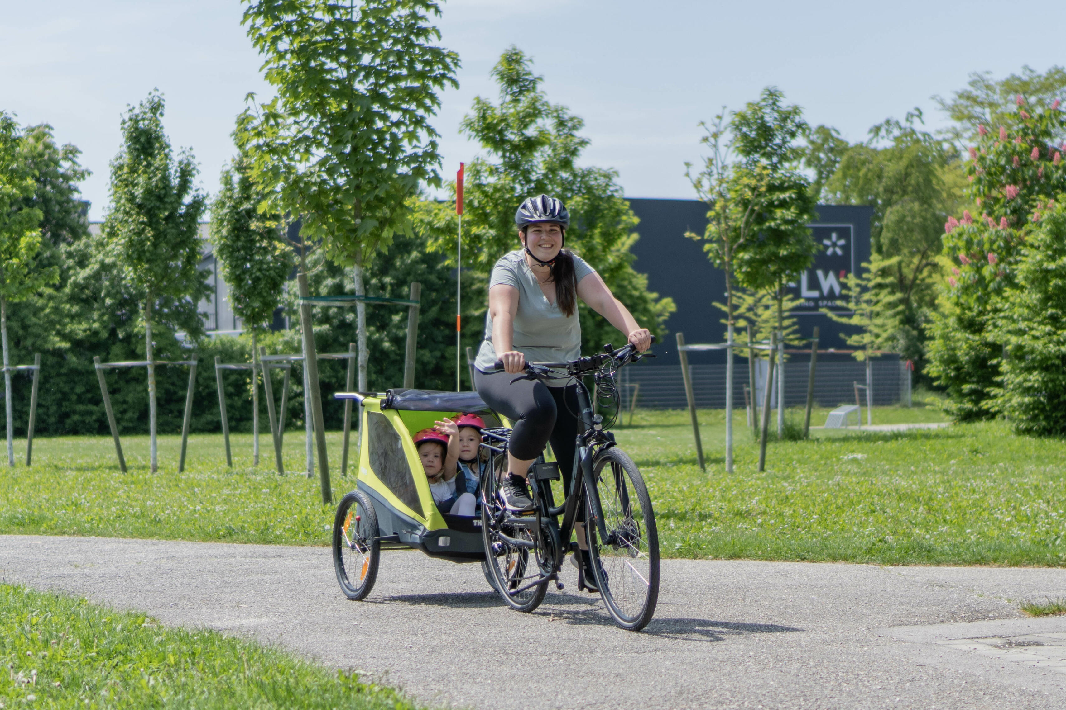 Dame fährt mit Fahrrad durch den Park. Im Anhänger sitzen 2 Mädchen mit pinken Fahrradhelmen.