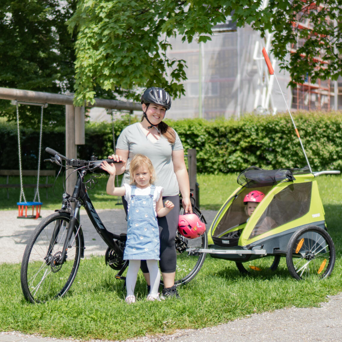 Dame mit 2 Kinder steht mit Fahrrad und Kinderanhänger auf Wiese bei Kinderspielplatz.