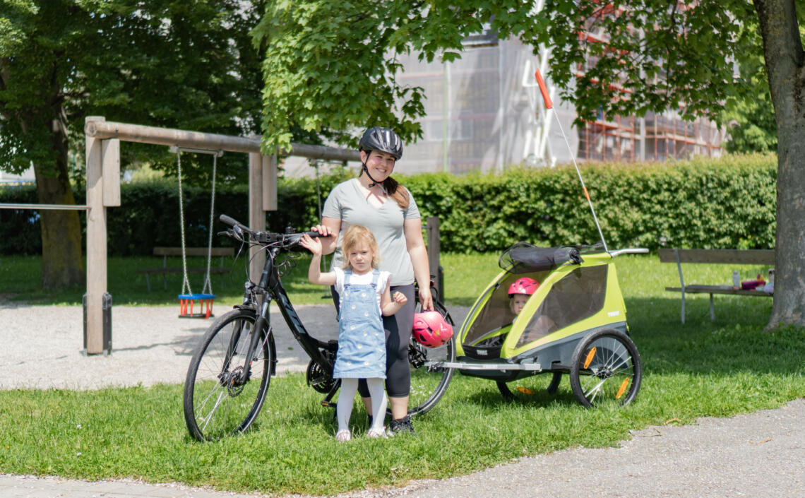 Dame mit 2 Kinder steht mit Fahrrad und Kinderanhänger auf Wiese bei Kinderspielplatz.