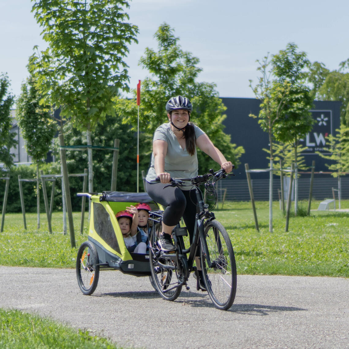 Dame fährt mit Fahrrad durch den Park. Im Anhänger sitzen 2 Mädchen mit pinken Fahrradhelmen.