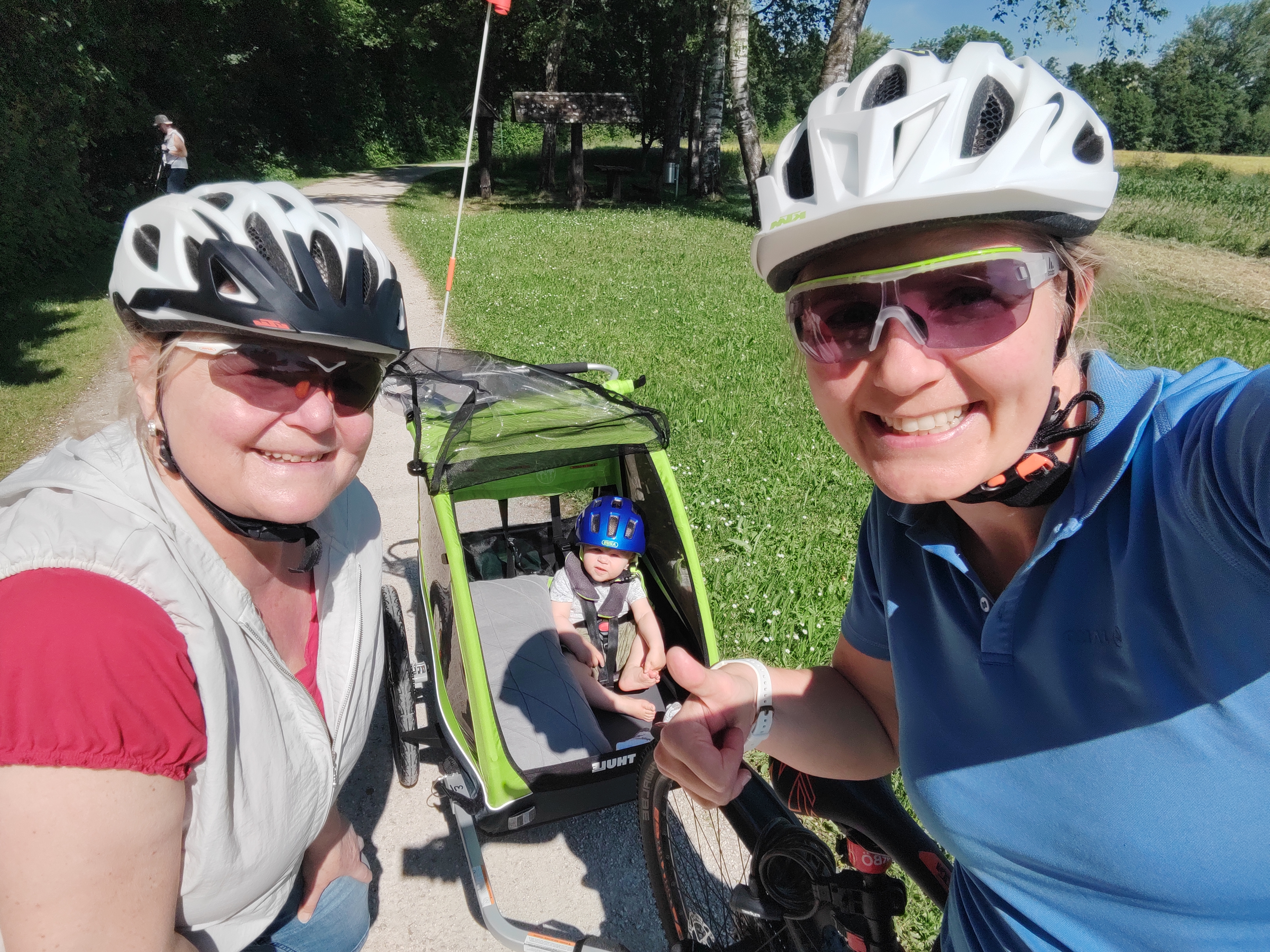 Zwei Frauen mit Fahrradhelm lachen, im Hintergrund Kind im Fahrradanhänger