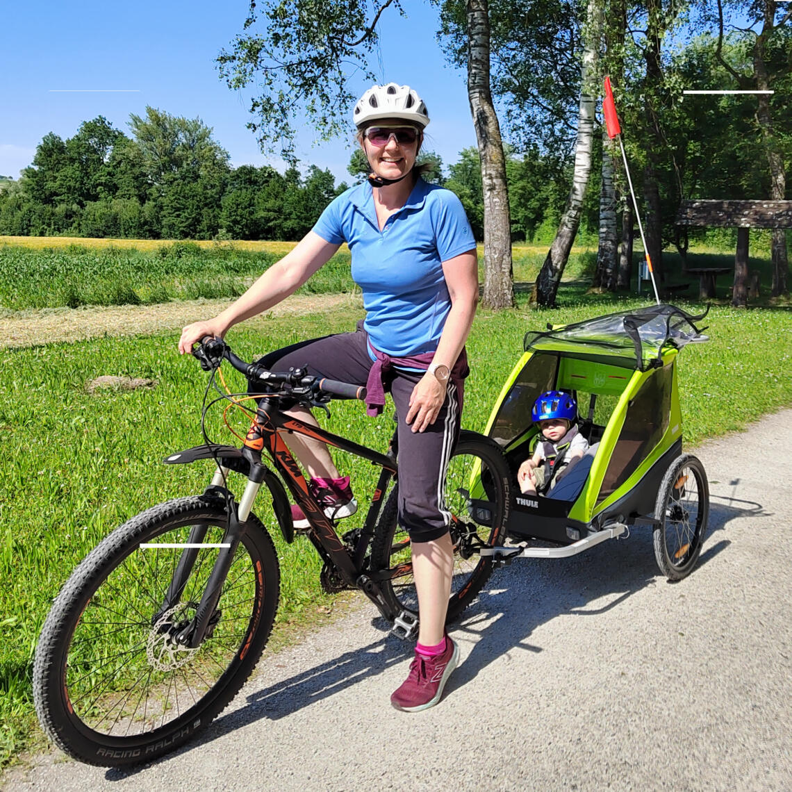 Dame mit Fahrrad und Kinderanhänger lächelt. Im Anhänger sitzt ein Kind mit blauen Fahrradhelm.