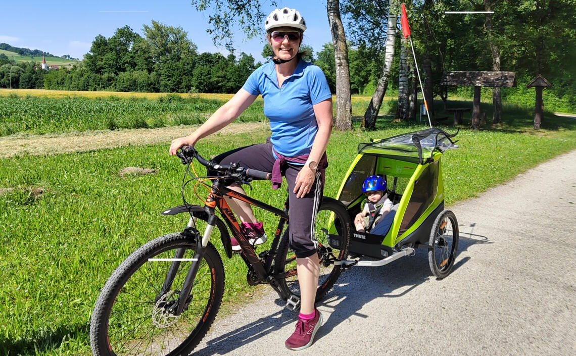 Dame mit Fahrrad und Kinderanhänger lächelt. Im Anhänger sitzt ein Kind mit blauen Fahrradhelm.