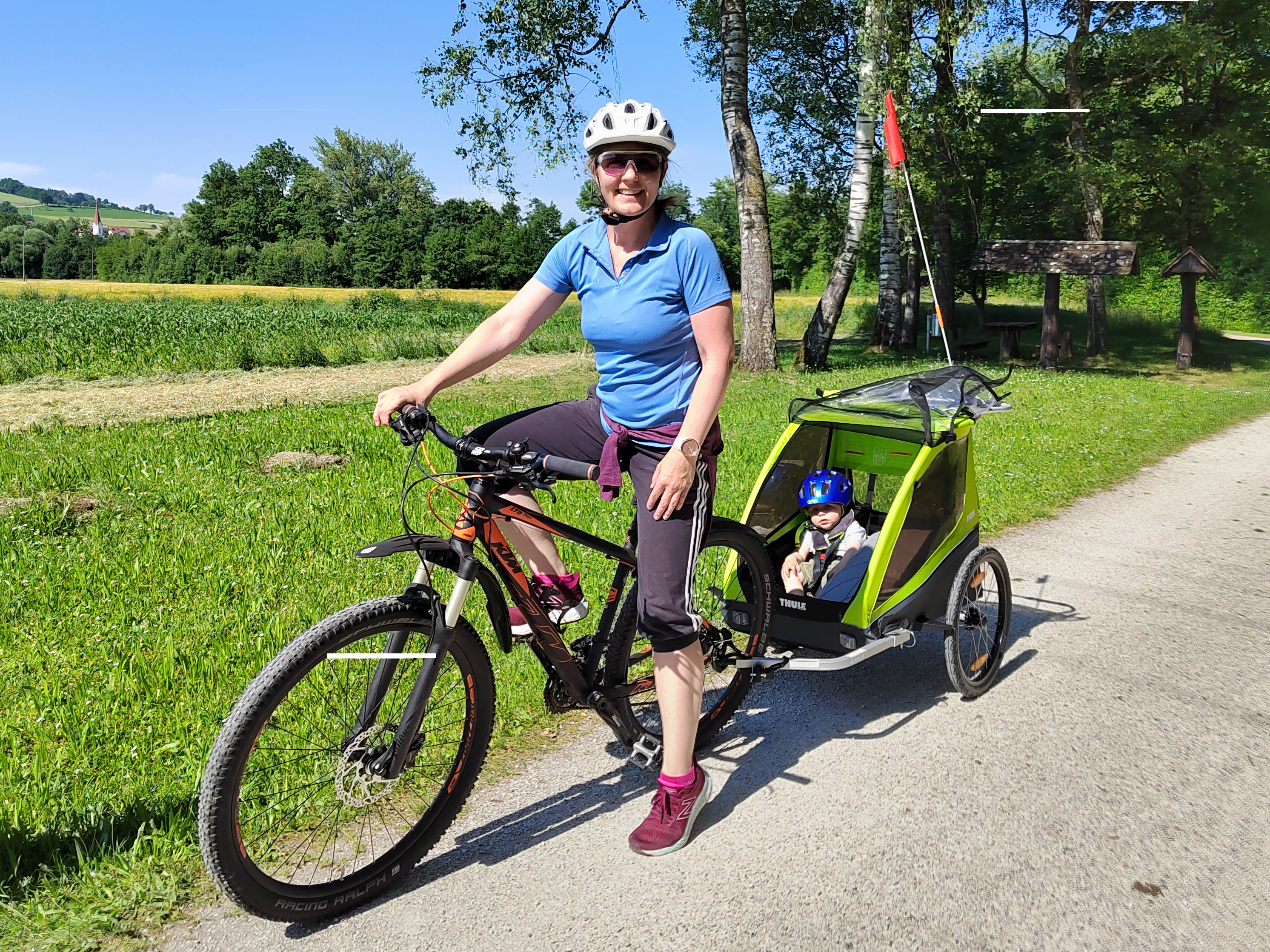 Dame mit Fahrrad und Kinderanhänger lächelt. Im Anhänger sitzt ein Kind mit blauen Fahrradhelm.