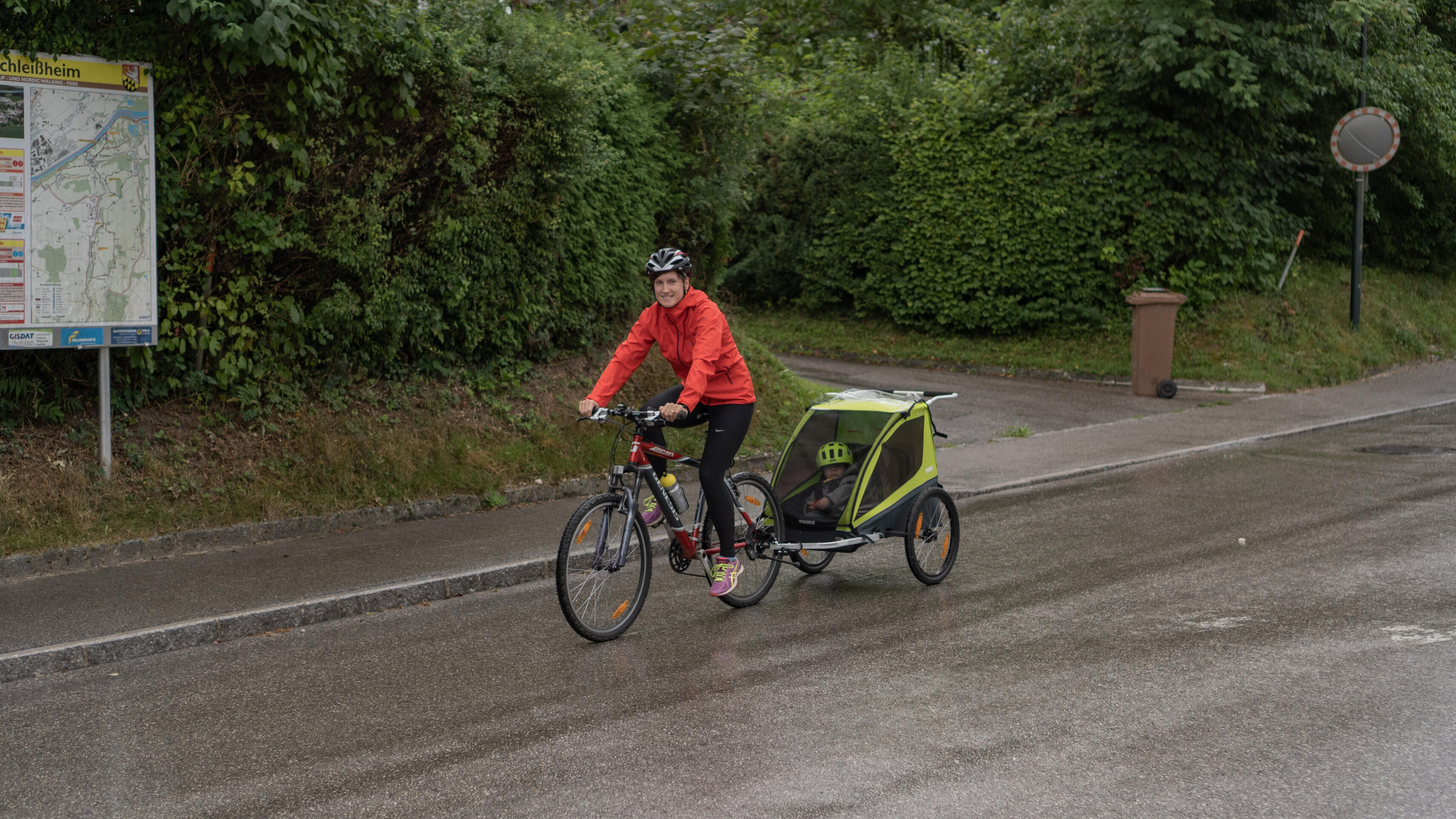 Dame fährt mit roter Regenjacke und Fahrrad mit Kinderanhänger auf einer nassen Straße