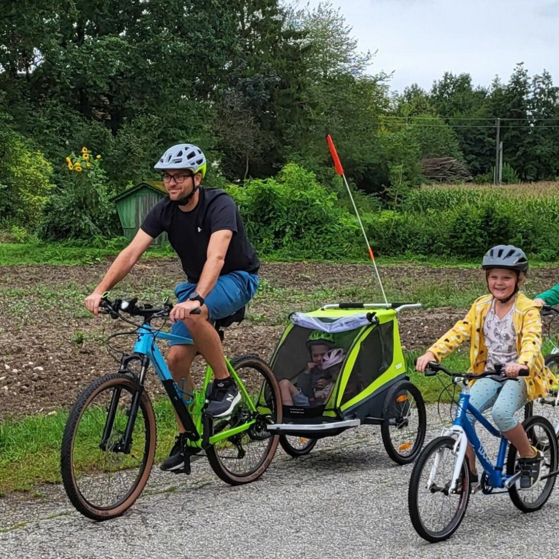 Vater zieht Kinderradanhänger mit zwei Kindern im Anhänger, zwei Kinder radeln selber, ländlicher Hintergrund mit Feldern und Bienenstöcken