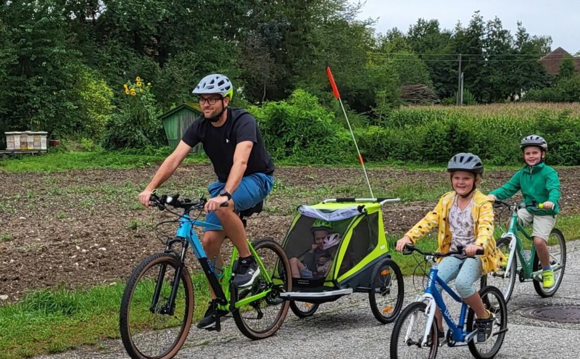 Vater zieht Kinderradanhänger mit zwei Kindern im Anhänger, zwei Kinder radeln selber, ländlicher Hintergrund mit Feldern und Bienenstöcken