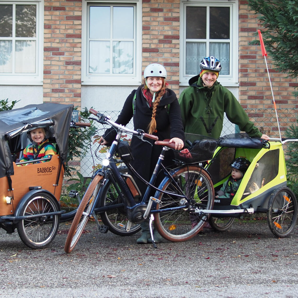 Foto von 4-köpfiger Familie, Mann und Frau mit Fahrrädern, beide haben einen Kinderradanhänger, in den Anhängern sitzen zwei Kinder, im Hintergrund ist eine Hausmauer