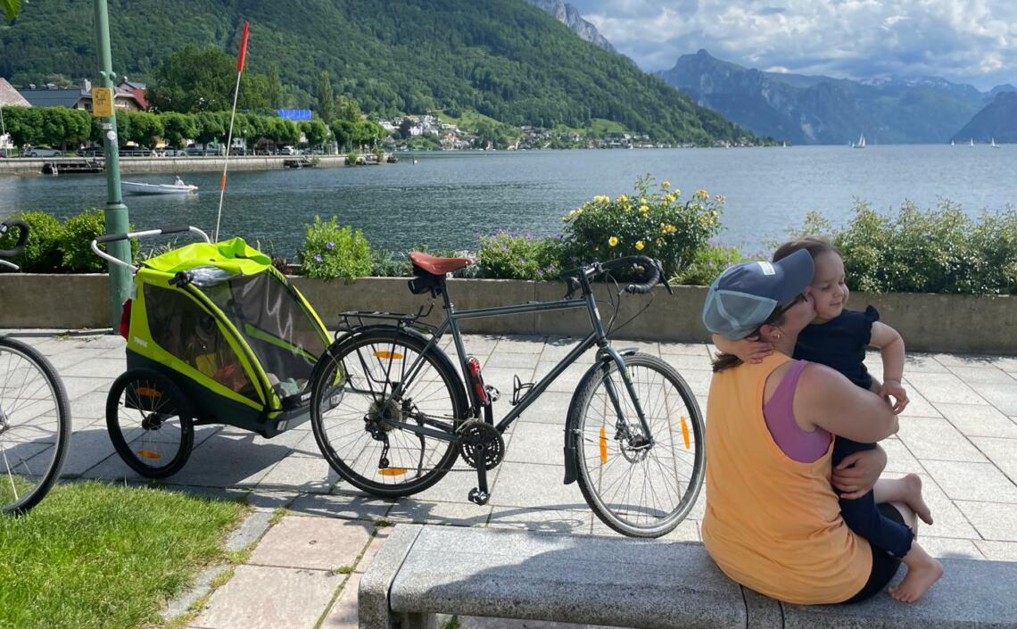 Mutter mit Tochter an einem See, mit Kinderradanhänger, Berge im Hintergrund