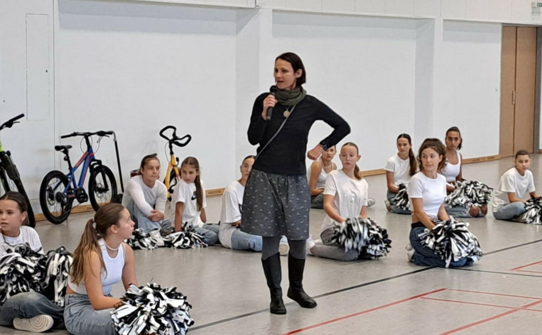 Frau spricht mit Mikrofon vor einer Gruppe von Cheerleadern im Turnsaal, an der Rückwand stehen drei Fahrräder