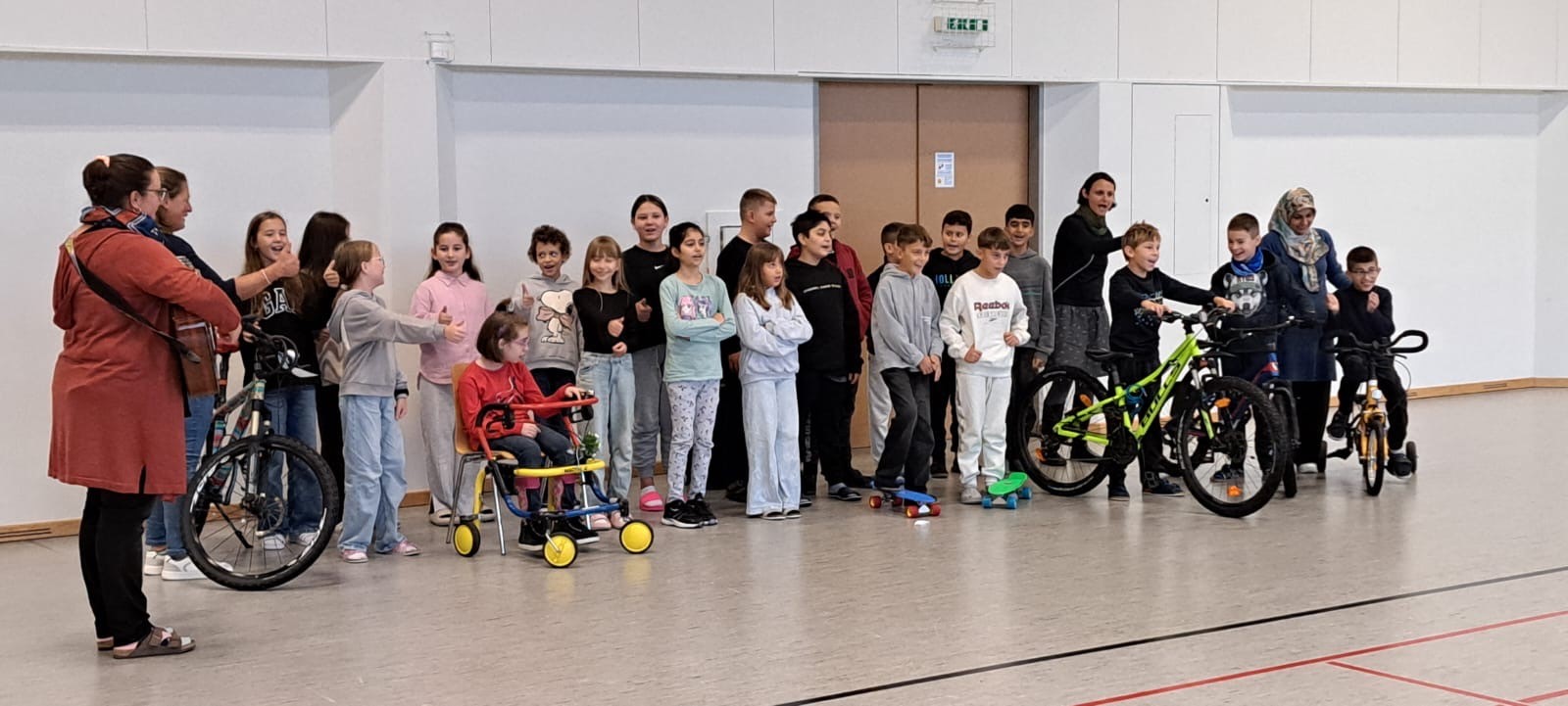 Schulklasse in einem Turnsaal teilweise mit Fahrrädern, Skateboards, Rollstuhl, Fahrrad mit Stützrädern