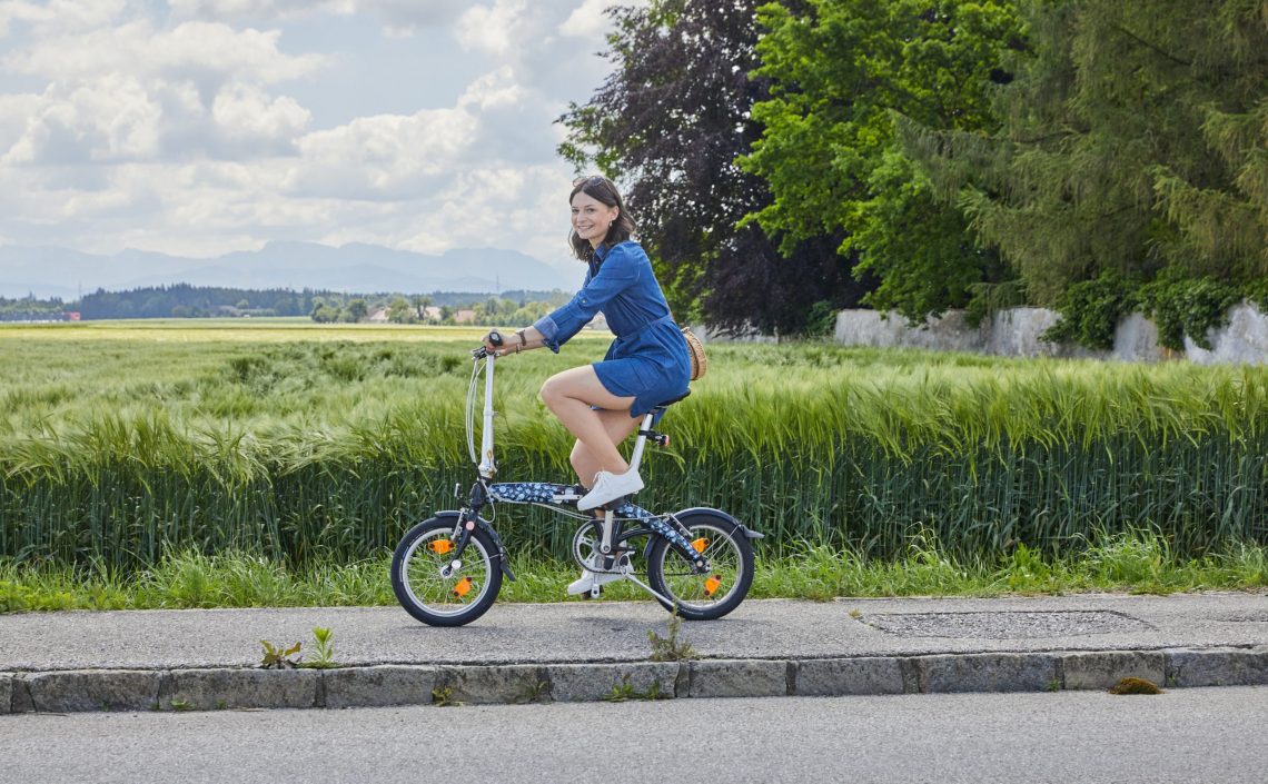 Frau auf Klapprad auf Gehsteig neben einem Feld